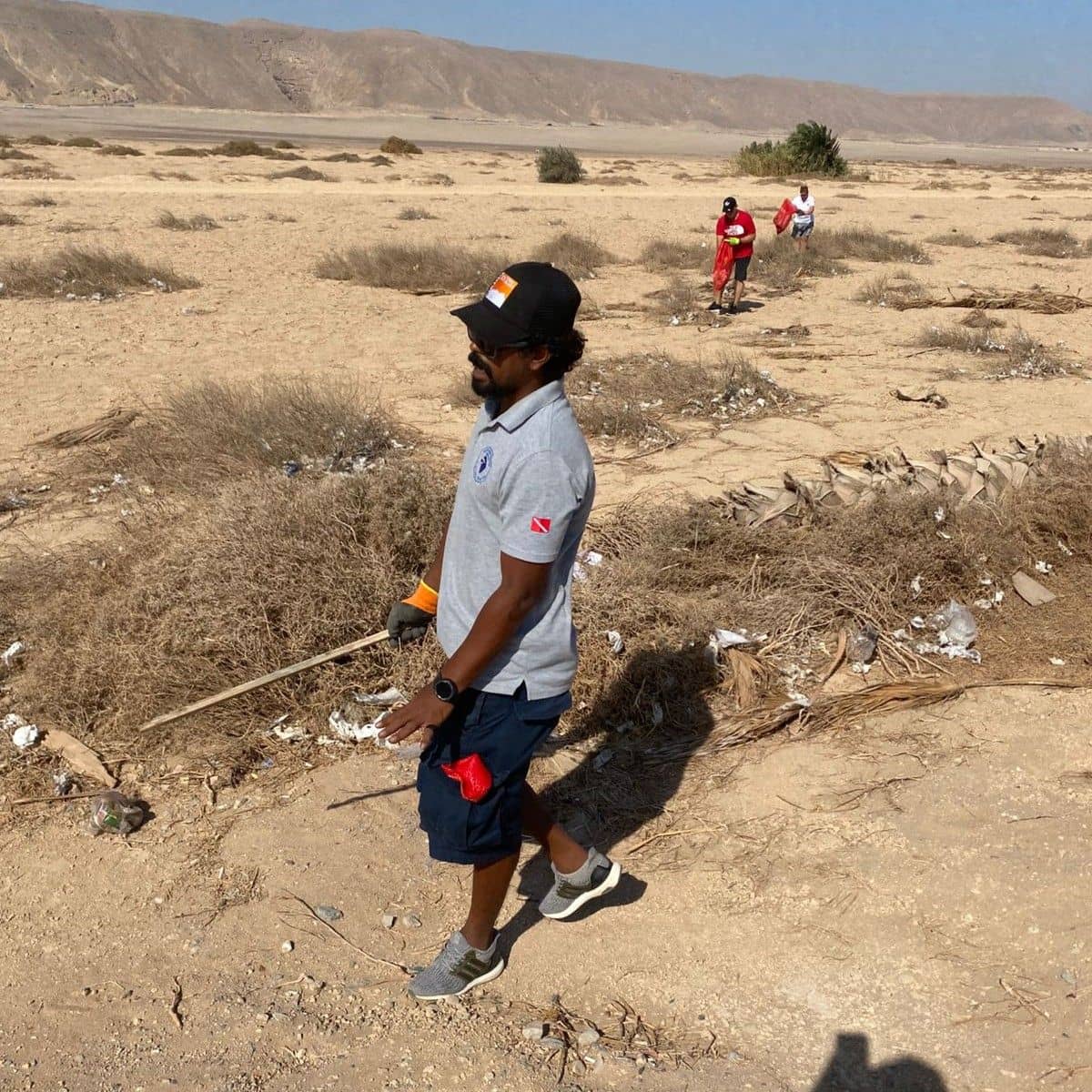 Man walks through a dry, sandy landscape holding a stick, with others in the background.