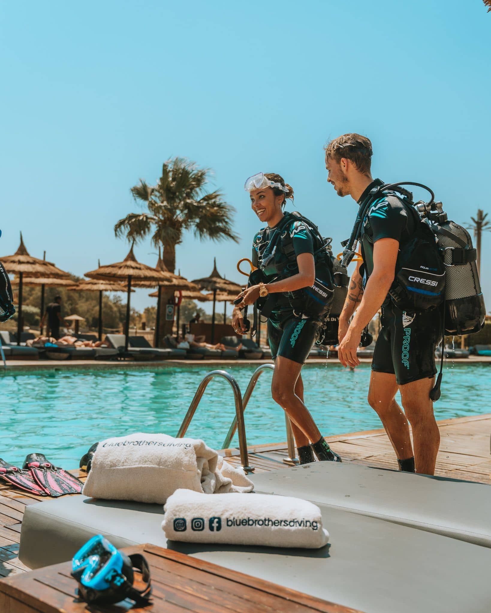 Two divers wearing full scuba gear walking into a shallow, clear blue swimming pool.