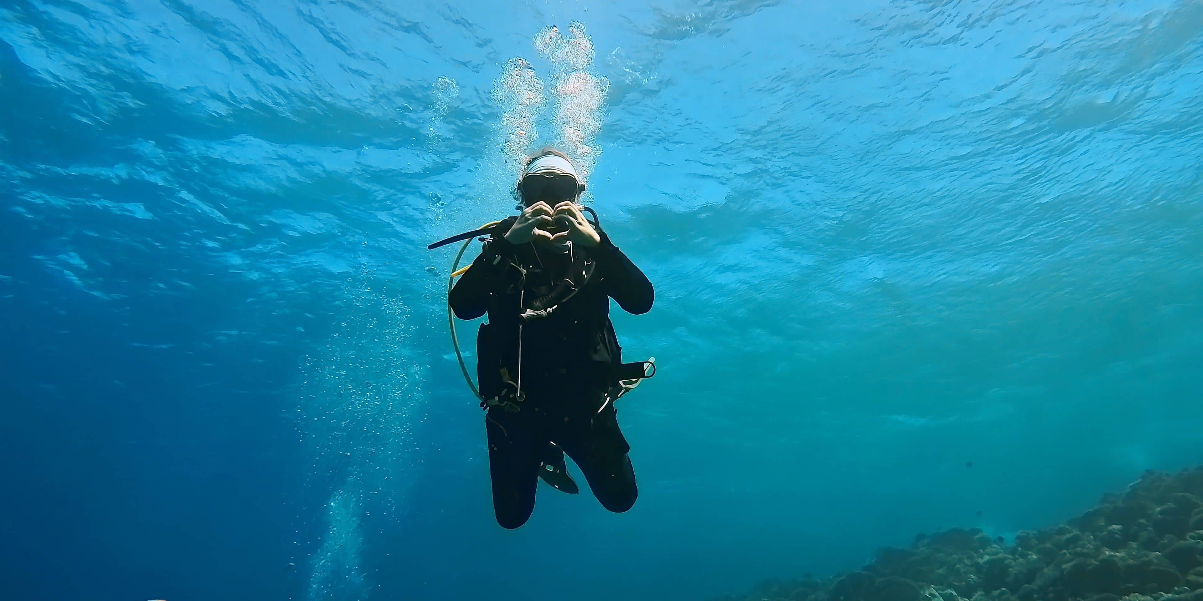 A scuba diver in a black wetsuit exhales bubbles underwater, with light filtering from the surface.