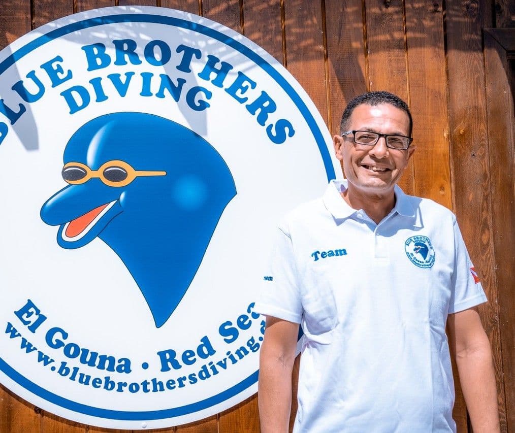 Kareem aus dem Blue Brothers Diving Team neben dem Schild der Tauchbasis