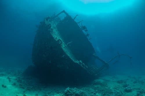 A large, decaying shipwreck resting on the sandy seabed, with sunlight illuminating the water.